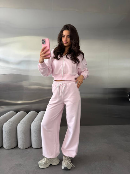 A woman takes a mirror selfie wearing a light pink two-piece tracksuit consisting of a cropped zip-up hoodie and matching wide-leg sweatpants, paired with gray sneakers.