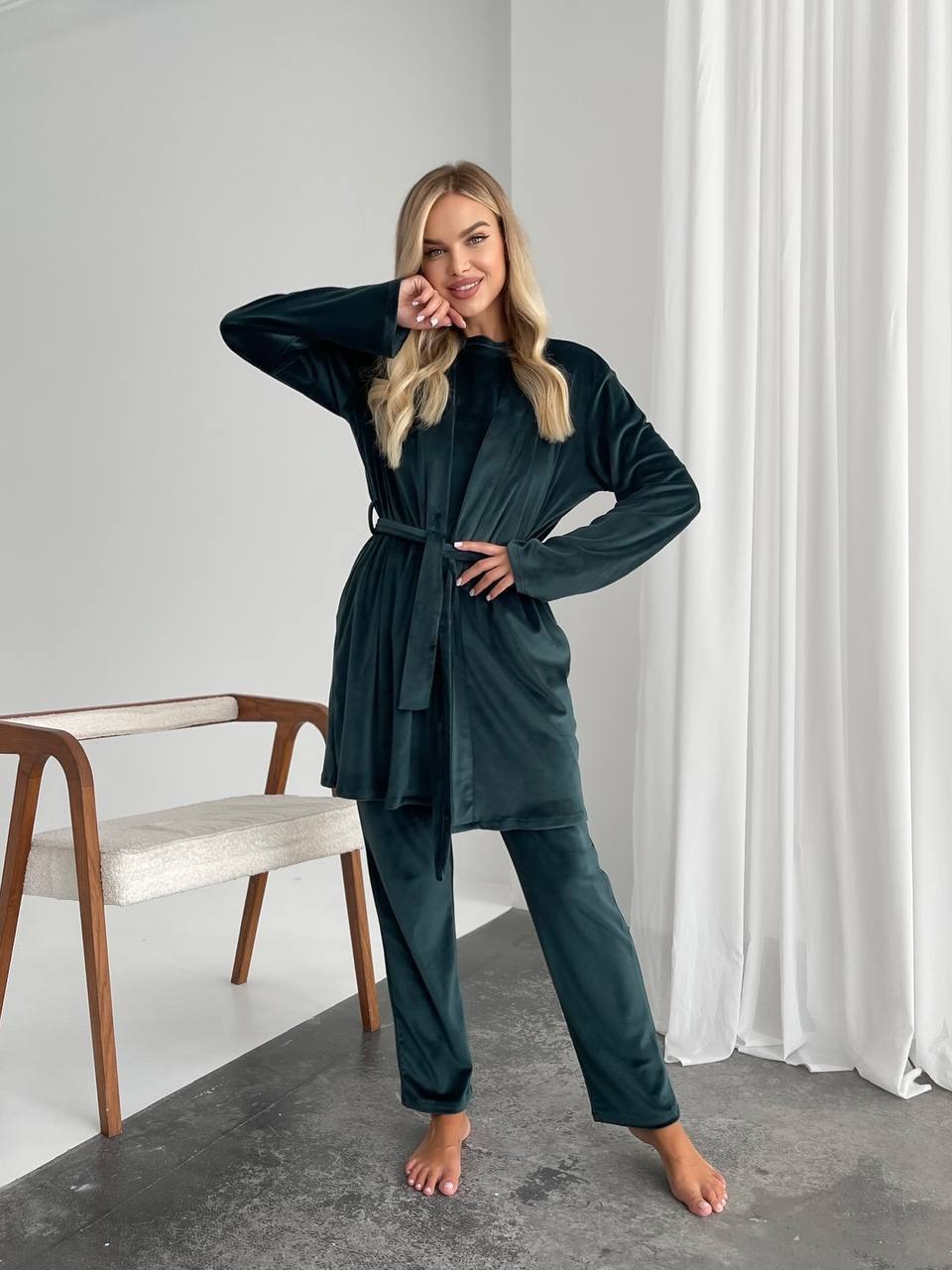 A woman modeling a dark teal two-piece loungewear set consisting of a long-sleeve belted robe and matching pants, standing in a minimalist room with white curtains and a wooden chair.