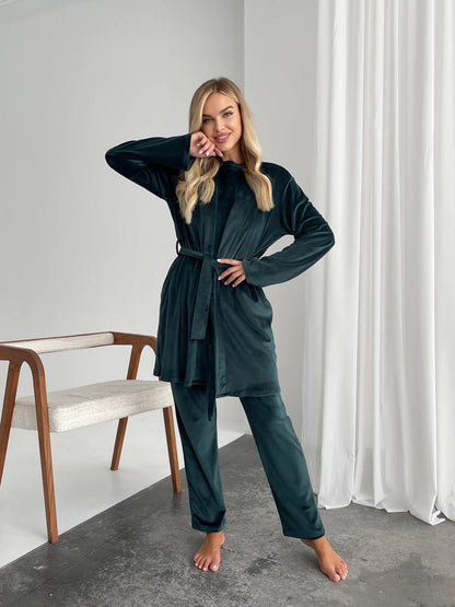 A woman modeling a dark teal two-piece loungewear set consisting of a long-sleeve belted robe and matching pants, standing in a minimalist room with white curtains and a wooden chair.