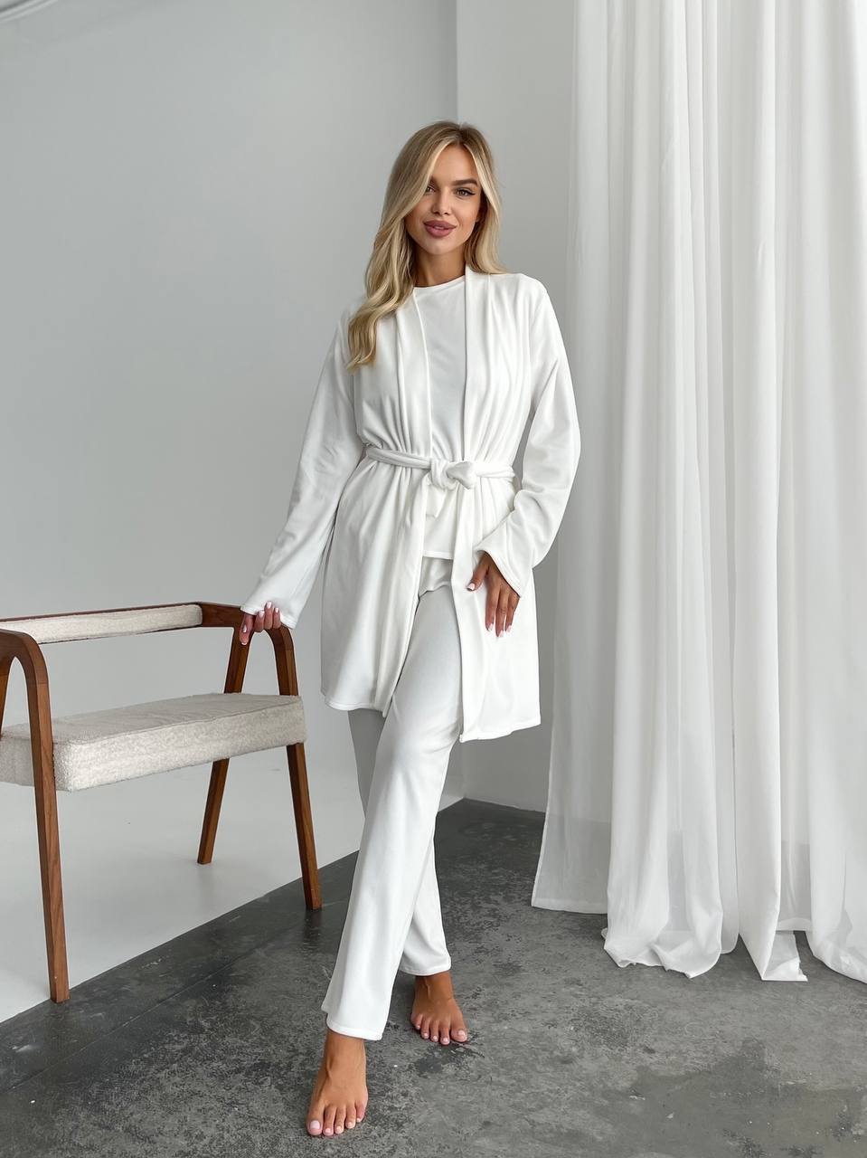 A woman models a white two-piece lounge set consisting of a long-sleeve belted robe and matching pants, standing in a minimalist room with white curtains, a gray floor, and a wooden chair.