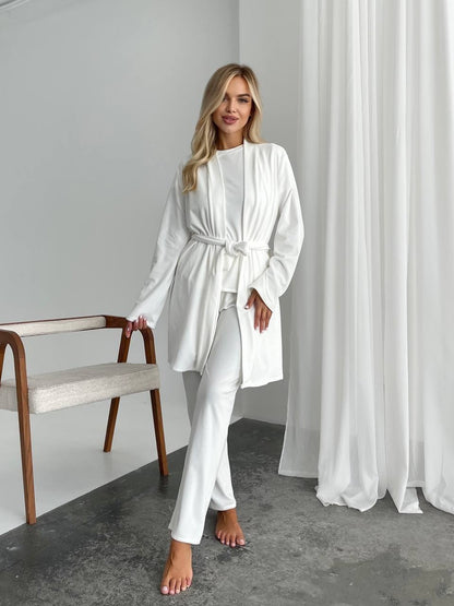 A woman models a white two-piece lounge set consisting of a long-sleeve belted robe and matching pants, standing in a minimalist room with white curtains, a gray floor, and a wooden chair.