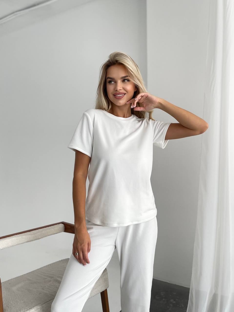 A woman posing in a minimalist white lounge set consisting of a short-sleeve crewneck t-shirt and matching wide-leg trousers, set against a bright, modern room with white walls and sheer curtains.