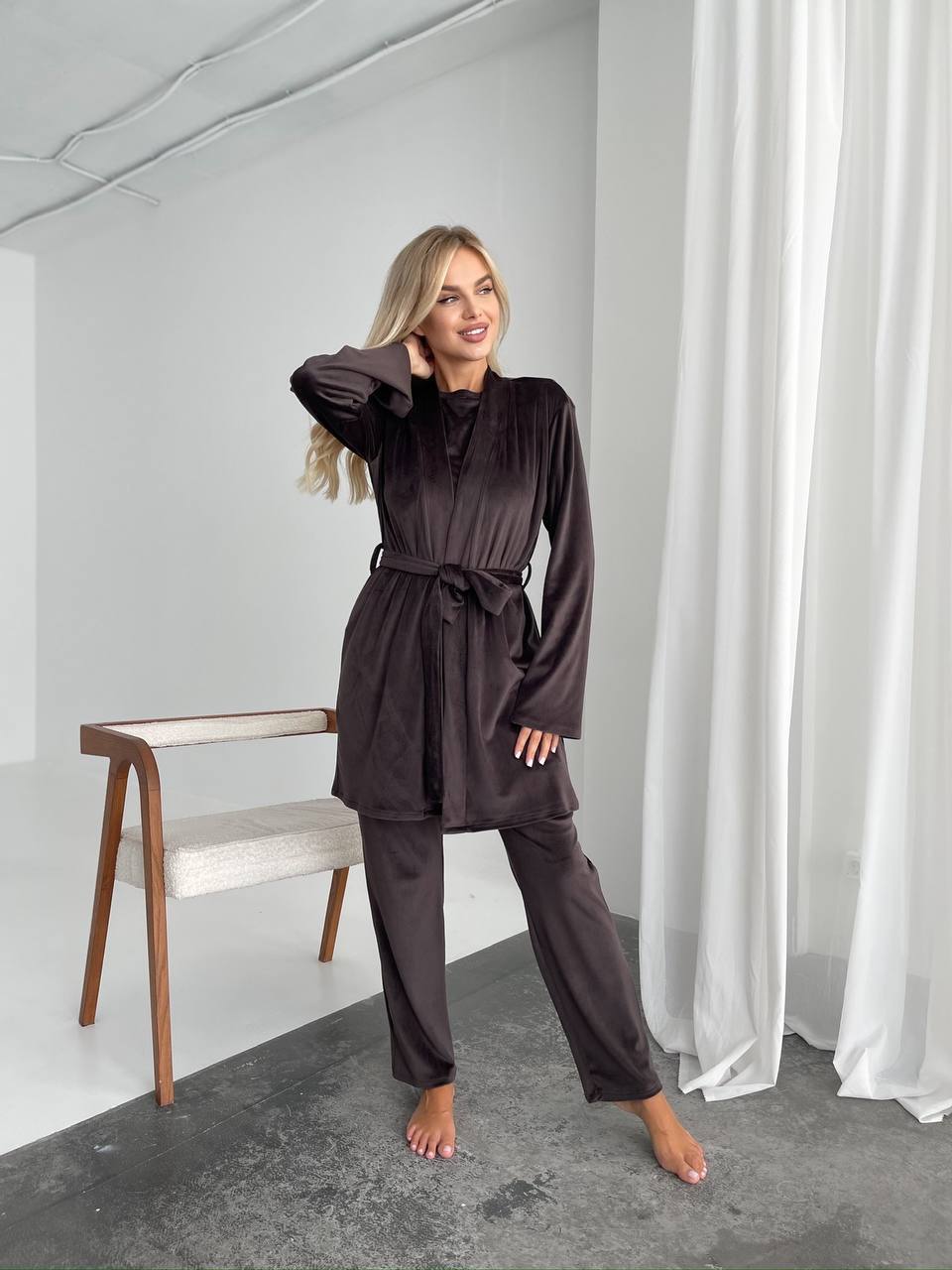 A woman modeling a dark brown two-piece lounge set consisting of a long-sleeve belted robe and matching pants, standing in a minimalist studio with white walls, a wooden chair, and white curtains.