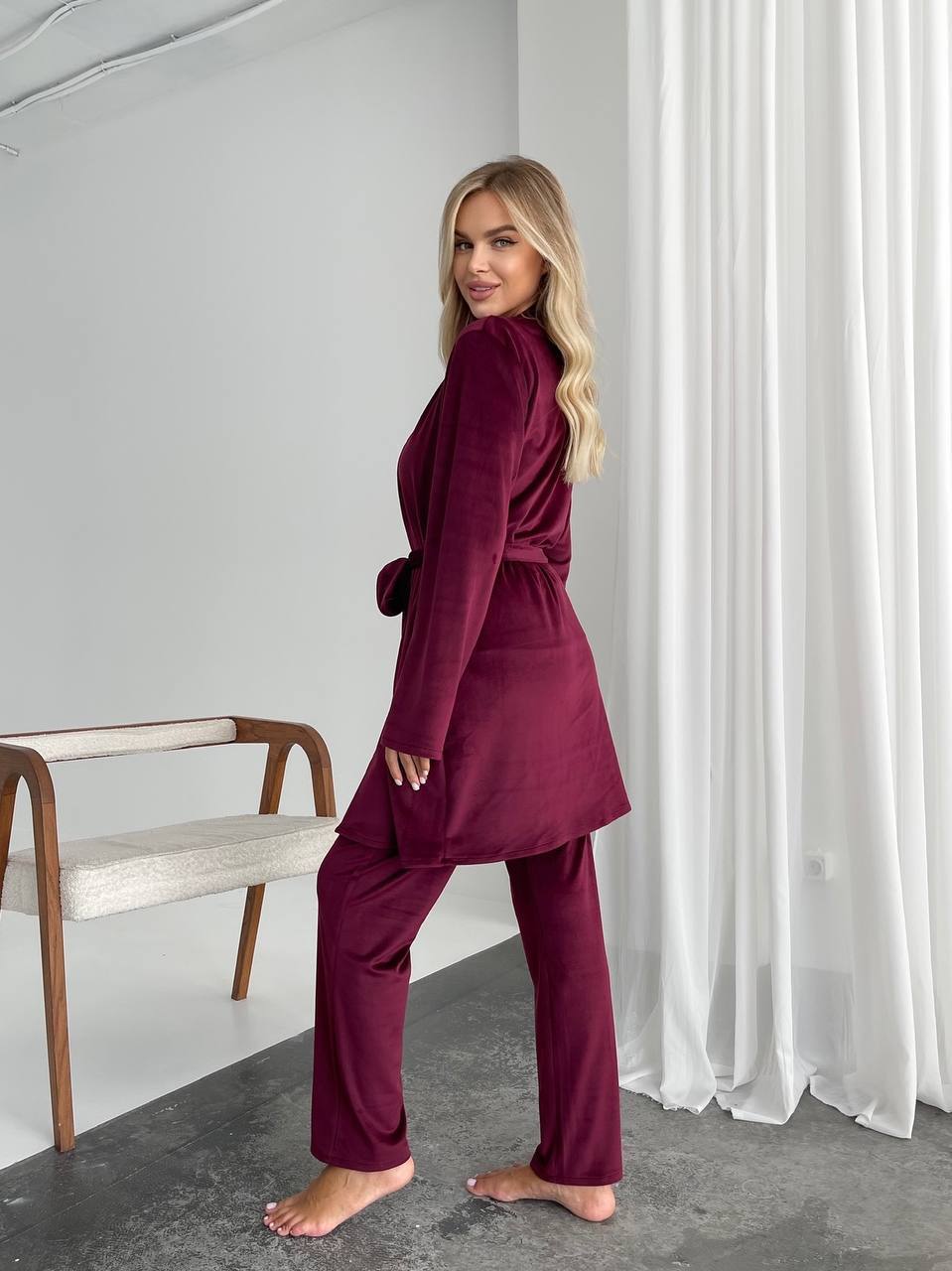 A woman modeling a matching two-piece lounge set in deep burgundy velvet, featuring a long-sleeve belted wrap top and matching wide-leg pants, standing barefoot in a minimalist room with white curtains.