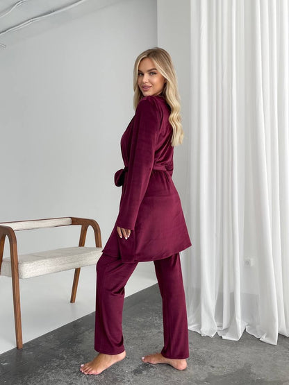 A woman modeling a matching two-piece lounge set in deep burgundy velvet, featuring a long-sleeve belted wrap top and matching wide-leg pants, standing barefoot in a minimalist room with white curtains.