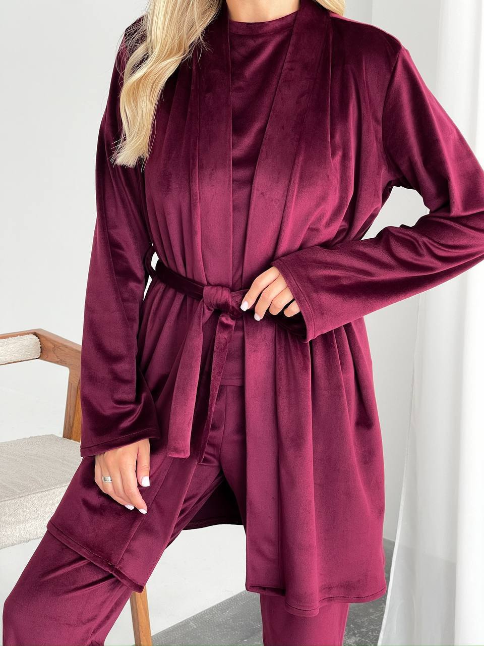 A woman wearing a matching burgundy velvet robe and pants set, standing indoors against a light neutral background.