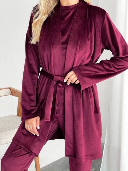 A woman wearing a matching burgundy velvet robe and pants set, standing indoors against a light neutral background.