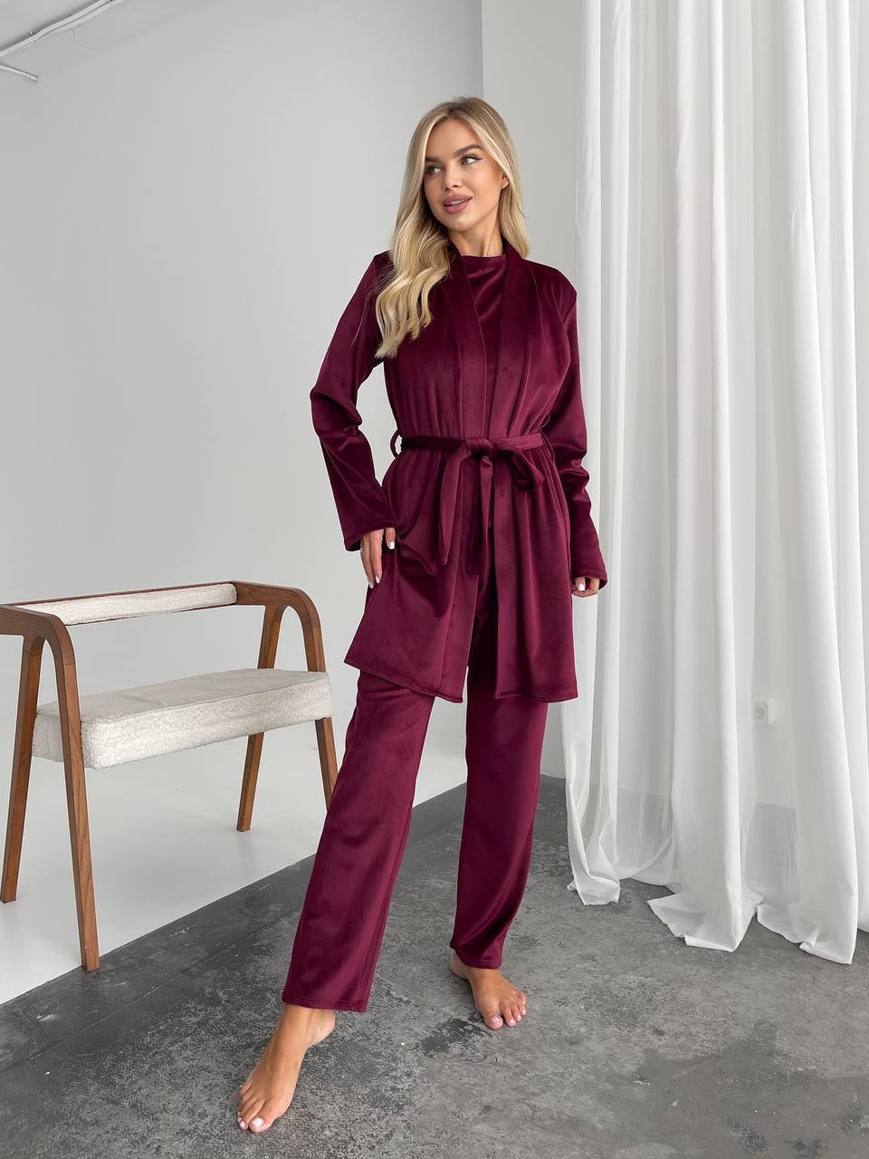 A woman modeling a matching burgundy velvet two-piece loungewear set consisting of a long-sleeve belted robe and wide-leg pants, standing in a minimalist room with a white curtain and a wooden chair.