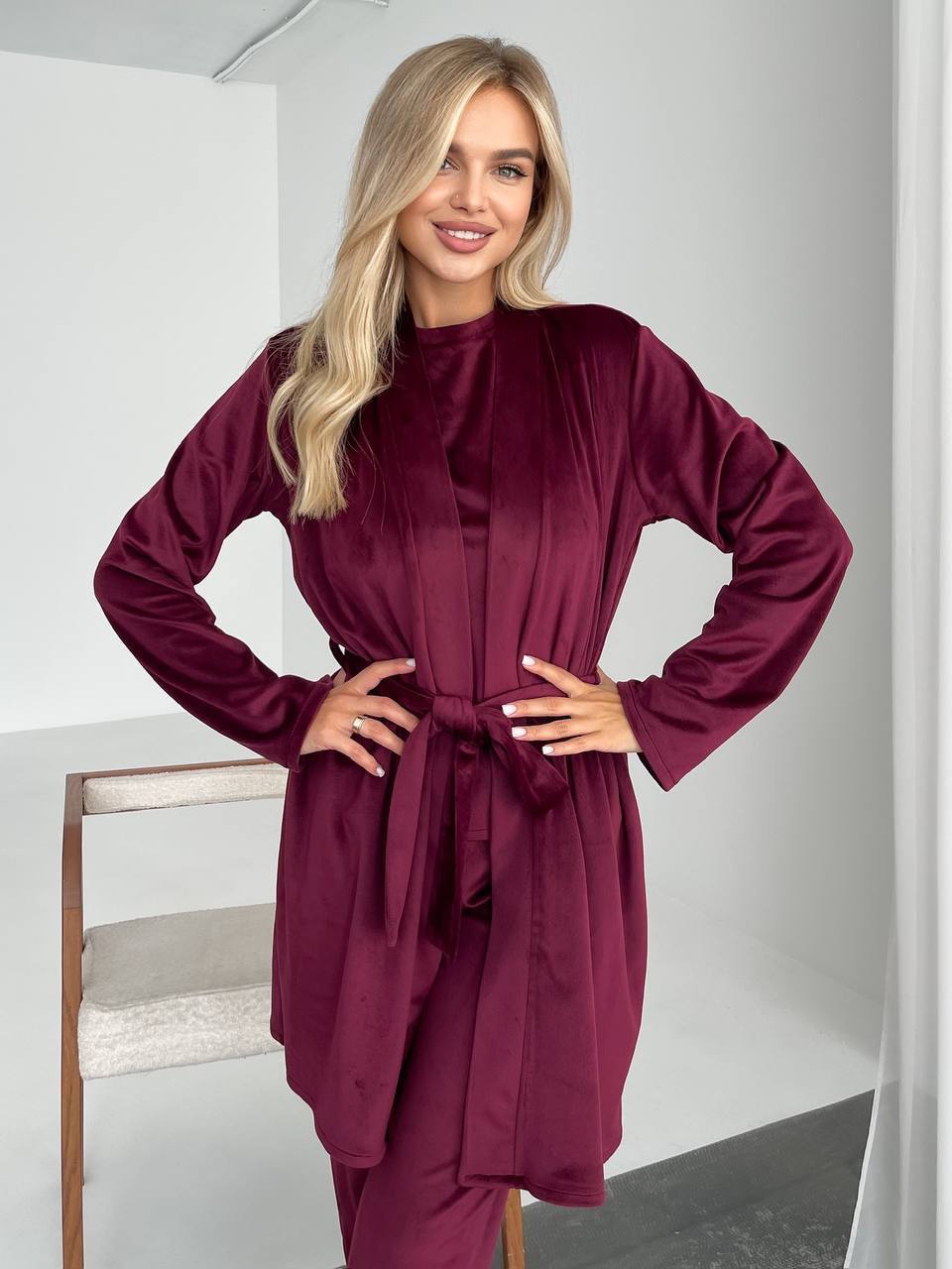 A woman modeling a long-sleeve, high-neck, belted robe in a deep burgundy velvet fabric, standing in a minimalist white room.