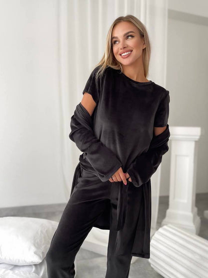 A woman wearing a black velvet lounge set consisting of a long-sleeve top with cutout sleeves and matching pants, standing in a bright, modern room with white columns.