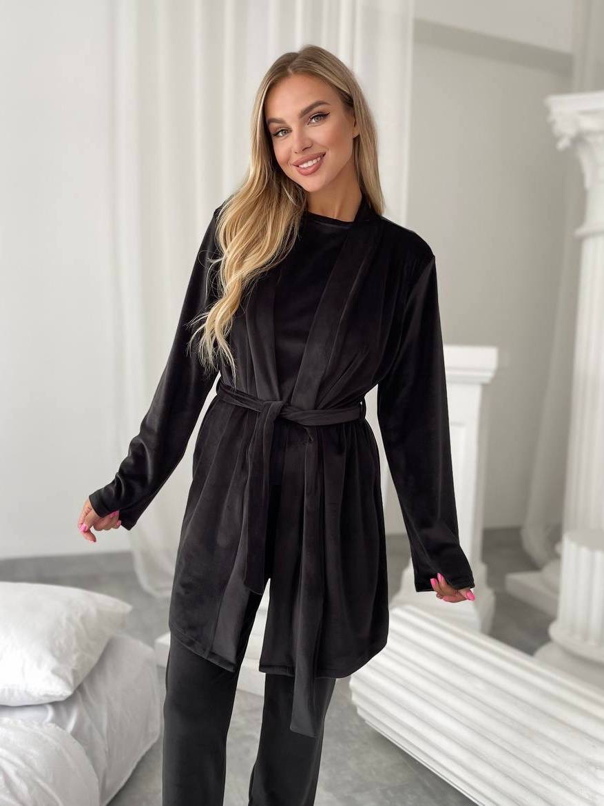A woman modeling a black velvet long-sleeve robe with a tie belt, paired with black pants, in a bright, modern bedroom setting with white columns and a bed.