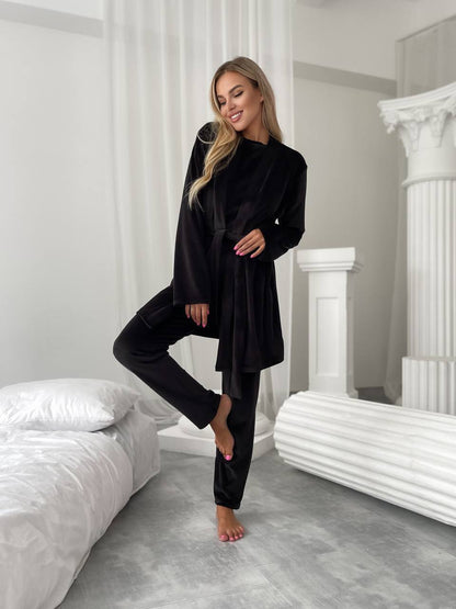 A woman modeling a black velvet lounge set consisting of a long-sleeve belted tunic top and matching pants, standing in a modern bedroom setting with white columns and a bed.