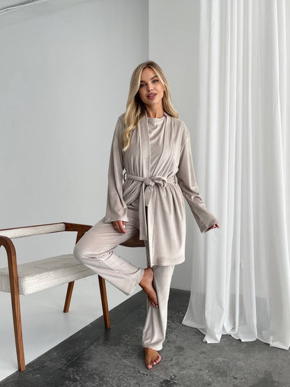 A woman models a matching two-piece loungewear set in a light taupe-beige satin-like fabric, consisting of a long-sleeve belted robe and matching wide-leg pants. She is posed in a minimalist room with white curtains, a gray floor, and a wooden bench.