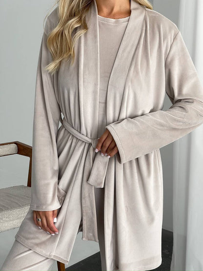A woman wearing a plush, long-sleeve beige robe with a matching belt and matching lounge pants, standing in a brightly lit indoor setting.