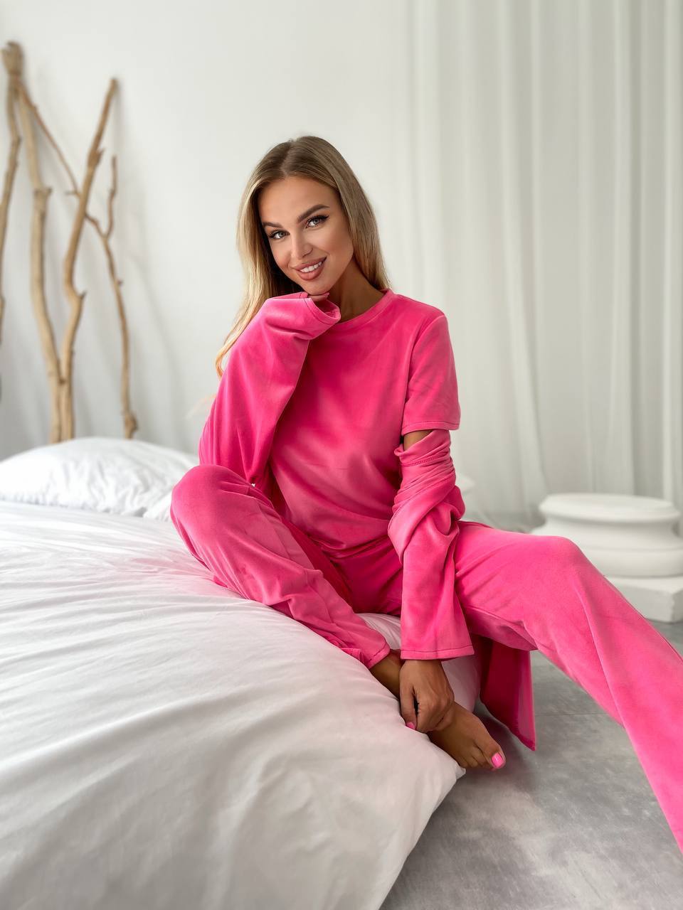 A woman wearing a bright pink lounge set with a matching top and pants, sitting on a white bed in a bright, modern bedroom.