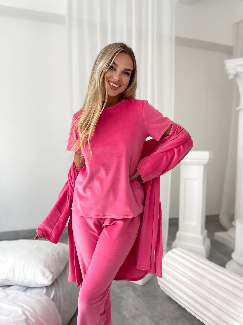 A woman modeling a bright pink lounge set consisting of a long-sleeve top and matching pants, with a matching pink cardigan draped over her shoulders, in a bright, modern bedroom setting.
