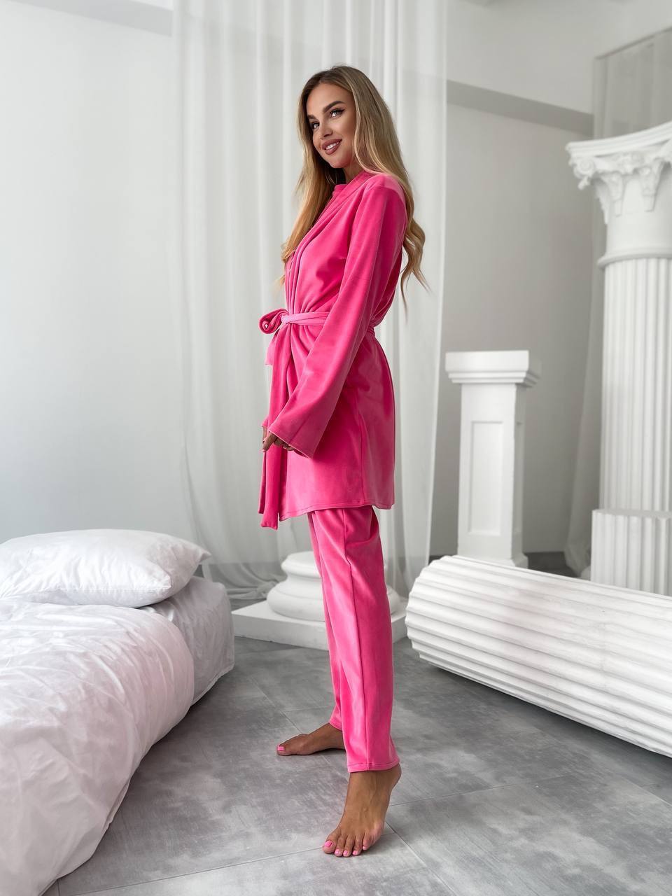 A woman modeling a bright pink velvet pajama set consisting of a long-sleeve belted robe and matching pants, standing in a modern, minimally decorated bedroom with white columns and a gray floor.