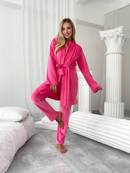 A woman modeling a bright pink two-piece loungewear set consisting of a long-sleeve belted robe and matching pants, standing in a modern white bedroom setting.