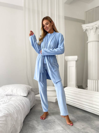 A woman modeling a light blue two-piece pajama set consisting of a long-sleeve robe and matching pants, standing in a bright bedroom with white bedding and classical-style columns.