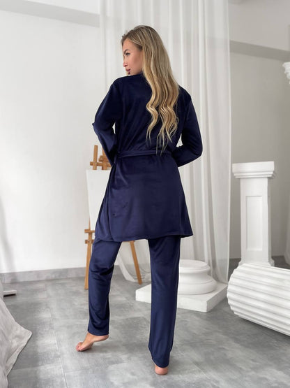 A woman wearing a navy blue two-piece pajama set, consisting of a long-sleeve belted robe and matching pants, standing barefoot in a bright, modern bedroom setting.