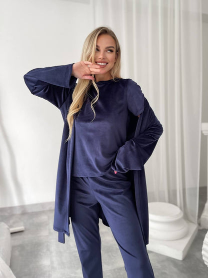 A woman modeling a matching navy blue loungewear set consisting of a long-sleeve top, matching pants, and a draped cardigan, set against a bright, modern indoor background with white curtains.