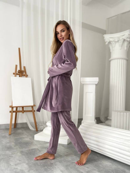 A woman modeling a matching two-piece pajama set in a bright, modern studio with white classical-style columns and an easel. The set consists of a long-sleeve belted robe and matching pants in a dusty lavender-purple velvet-like fabric.