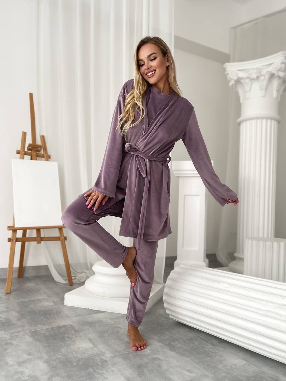 A woman modeling a matching two-piece lounge set in a bright, modern studio with white columns and gray flooring. The set consists of a long-sleeve wrap-style robe and matching pants in a dusty mauve-purple color.