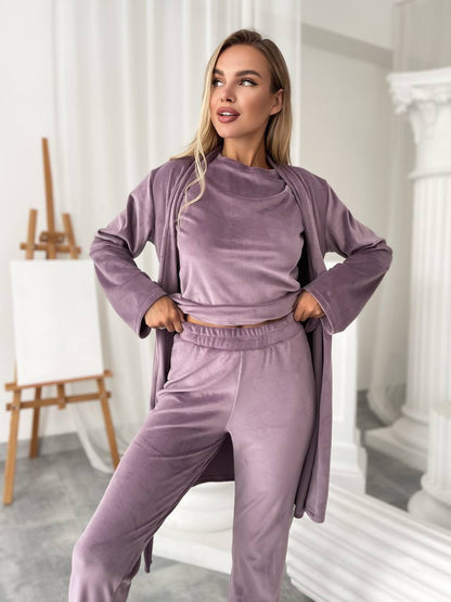 A woman modeling a matching lavender-purple velour lounge set consisting of a cropped long-sleeve top, jogger pants, and a draped cardigan, set against a bright, modern studio background with white columns and an easel.