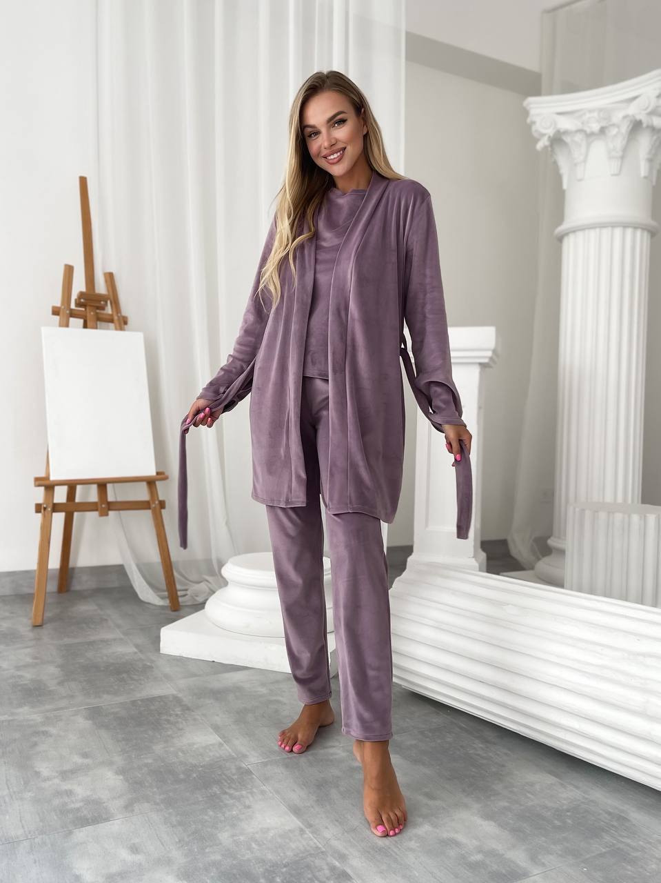 A woman modeling a matching two-piece pajama set in a bright, classical-style studio with white columns and an easel. The set consists of a long-sleeve button-front top and matching pants in a dusty mauve-purple velvet-like fabric.