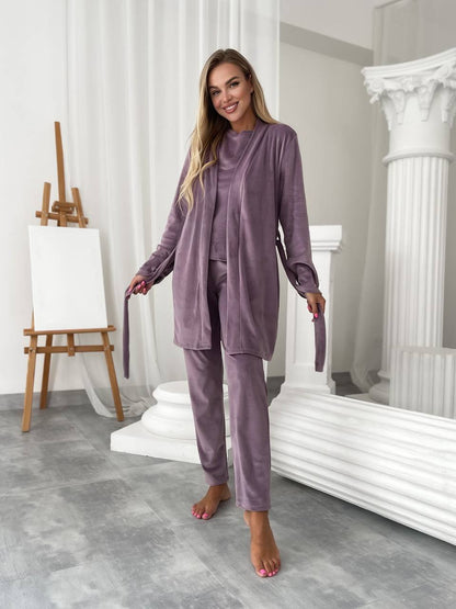 A woman modeling a matching two-piece pajama set in a bright, classical-style studio with white columns and an easel. The set consists of a long-sleeve button-front top and matching pants in a dusty mauve-purple velvet-like fabric.