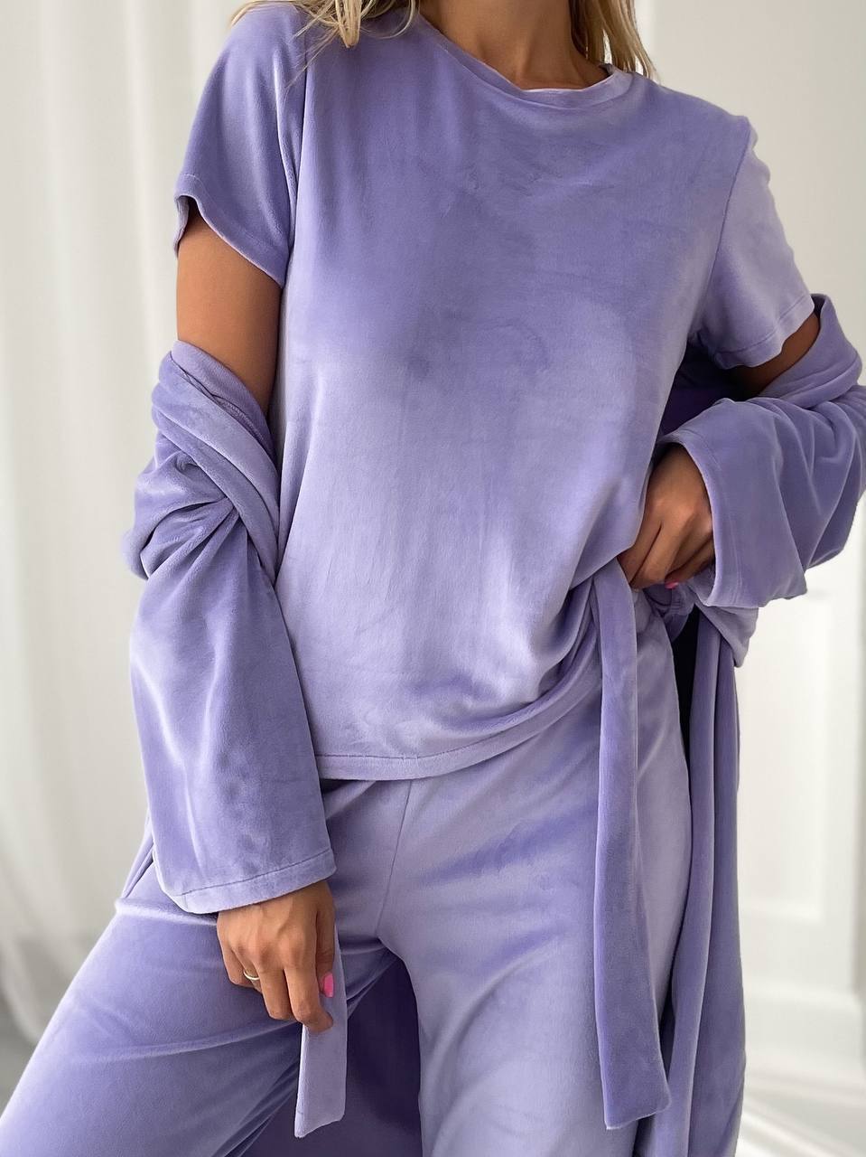 A woman wearing a lavender-purple velour lounge set consisting of a short-sleeve top with cutout sleeves and a matching long tie-front robe, paired with matching pants.