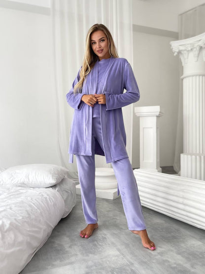 A woman modeling a matching lavender pajama set consisting of a long-sleeve button-front robe and coordinating pants, standing in a bright bedroom with white bedding and architectural columns.