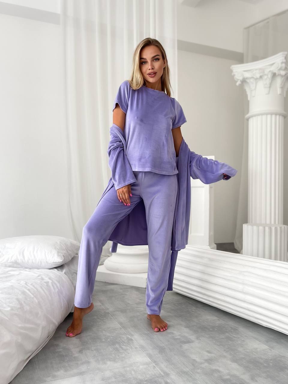 A woman models a lavender two-piece lounge set consisting of a short-sleeve cropped top with cutout details and matching sweatpants, styled with a matching zip-up cardigan in a bright, modern bedroom setting.