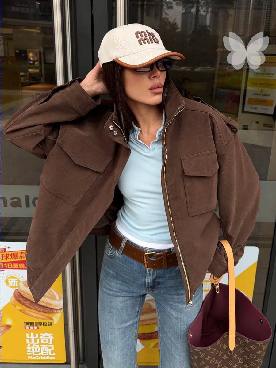 A woman wearing an oversized brown utility jacket over a light blue top and jeans, with a beige cap and sunglasses, holding a brown monogram handbag in front of a storefront.
