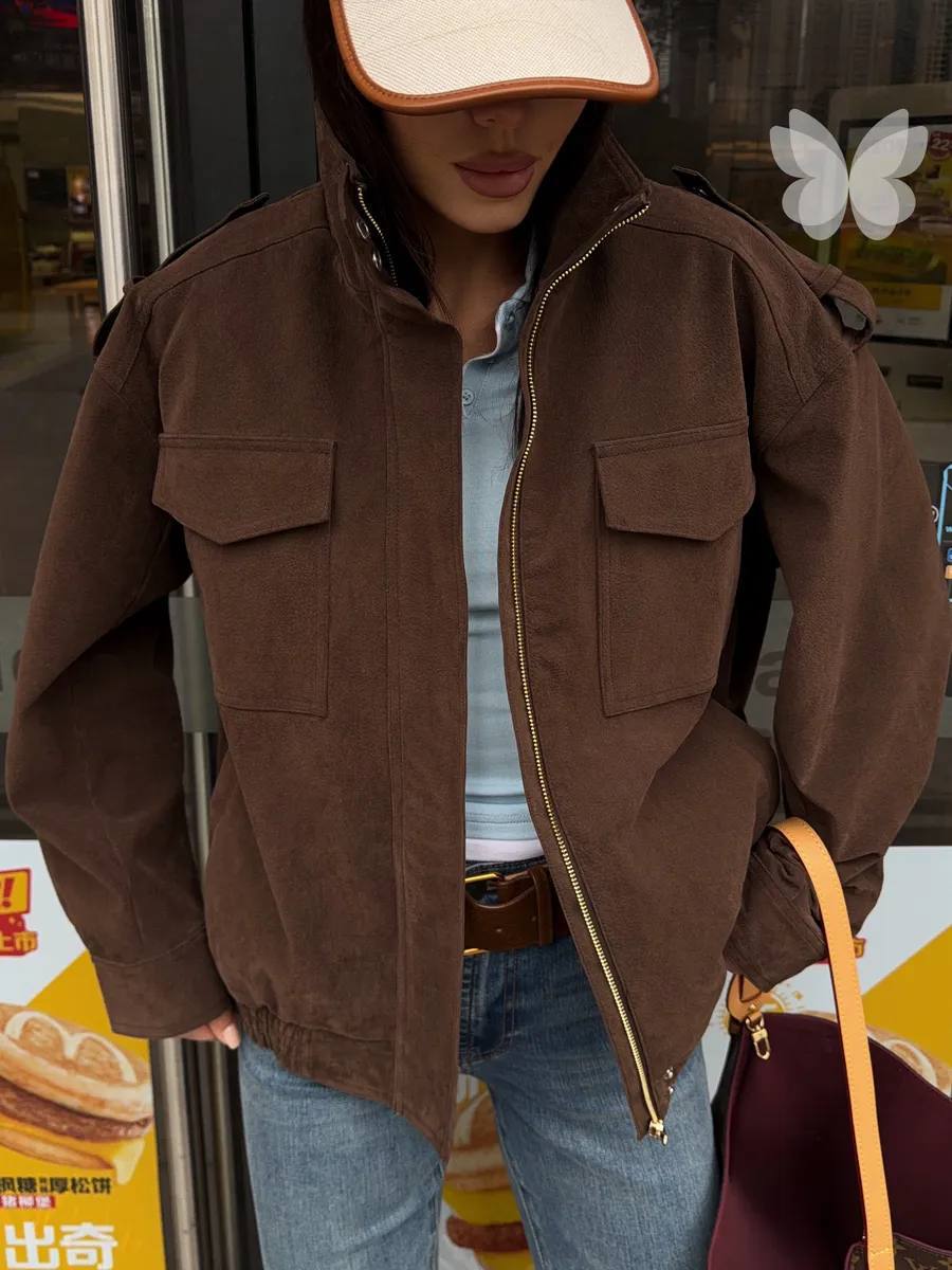 A person wearing a brown zip-front jacket with flap chest pockets, layered over a light blue top and blue jeans, standing in front of a storefront. A white visor and a burgundy shoulder bag are also visible.