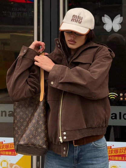 A person wearing a brown jacket, blue jeans, a beige Miu Miu cap, and holding a brown Louis Vuitton monogram shoulder bag in front of a storefront window.