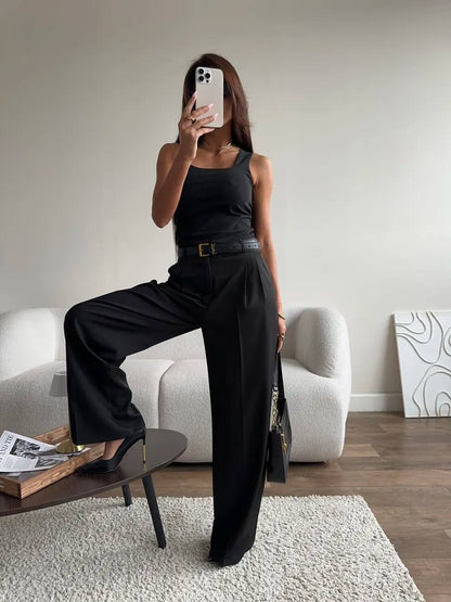 A woman takes a mirror selfie in a minimalist living room, wearing a black sleeveless top and black wide-leg trousers with a thin black belt. She is standing on a white textured rug next to a dark coffee table and a white sofa.