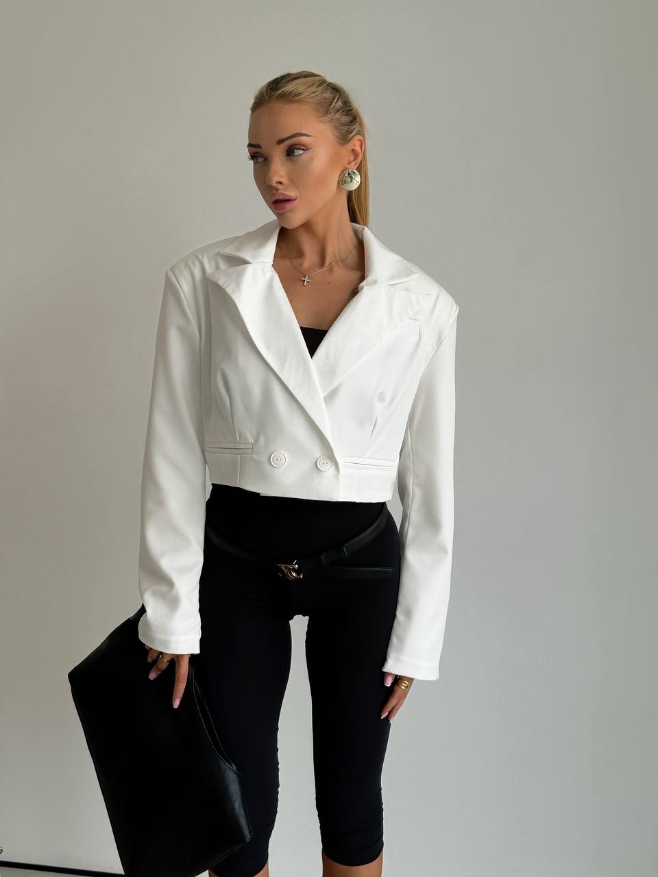A woman modeling a white cropped blazer with structured shoulders and double-breasted buttons, paired with black capri pants and holding a black clutch against a plain light gray studio background.