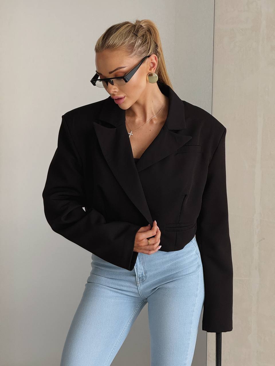 A woman wearing a black cropped blazer and light blue jeans, posing against a neutral beige wall.