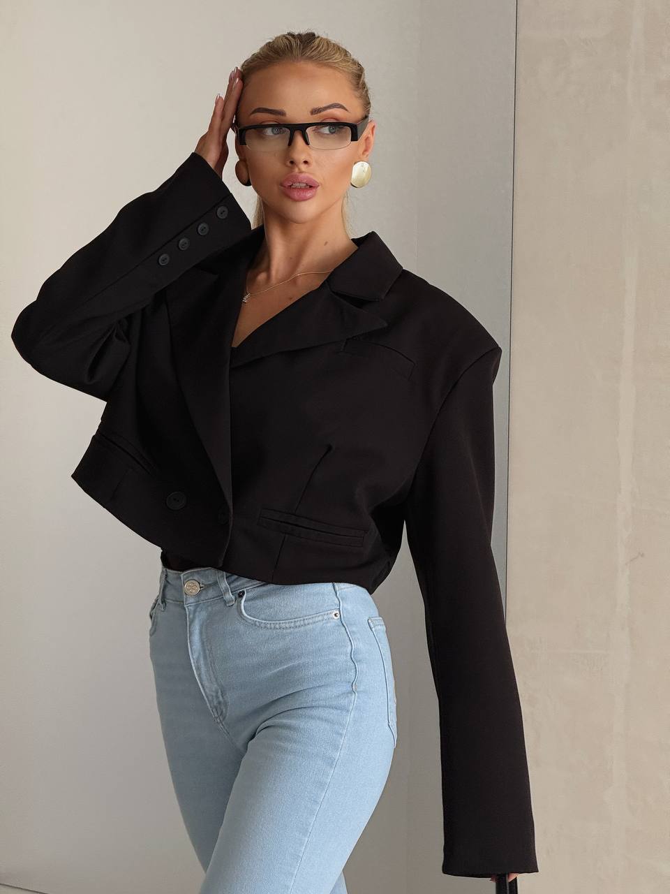 A woman wearing a black cropped blazer and light blue jeans, posing against a neutral beige wall.