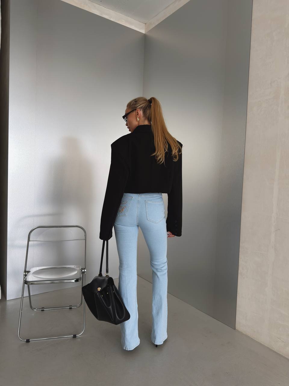 A woman stands with her back to the camera in a minimalist gray room, wearing a black cropped long-sleeve top and light blue flared jeans, holding a black handbag.