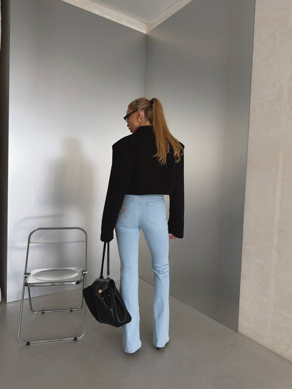 A woman stands with her back to the camera in a minimalist gray room, wearing a black cropped long-sleeve top and light blue flared jeans, holding a black handbag.