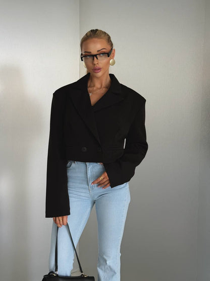 A woman modeling a black cropped blazer with wide lapels and long sleeves, paired with light blue jeans, standing against a plain light gray wall.