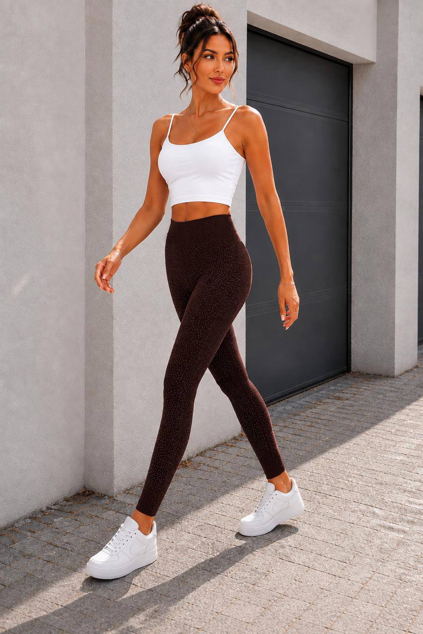 A woman modeling a matching activewear set consisting of a white spaghetti-strap crop top and dark brown textured leggings, paired with white sneakers, walking outdoors against a modern concrete wall and dark garage door.