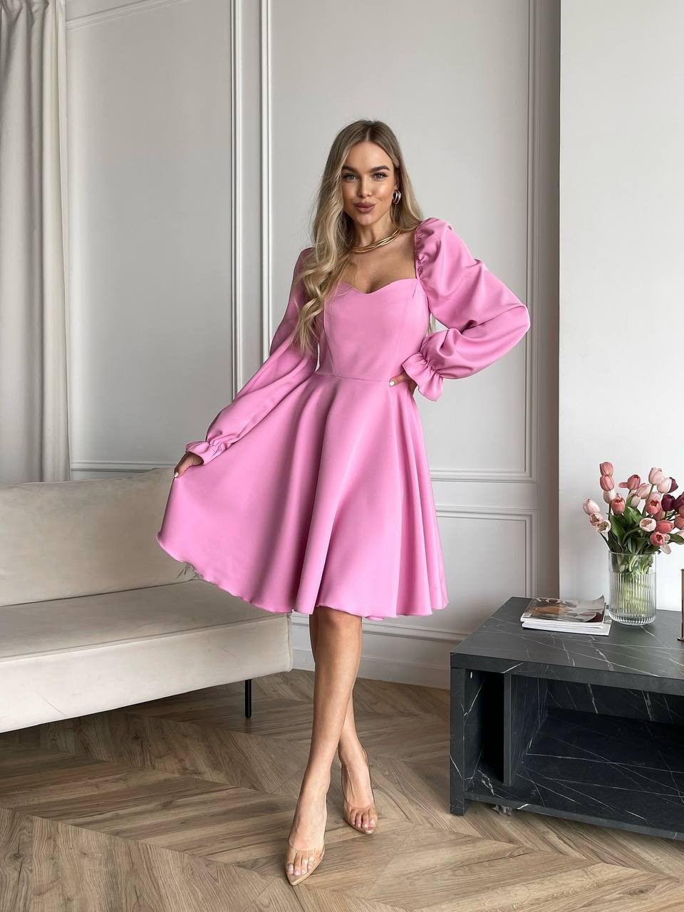 Woman wearing a pink dress in an elegant room with a sofa and decorative flowers.
