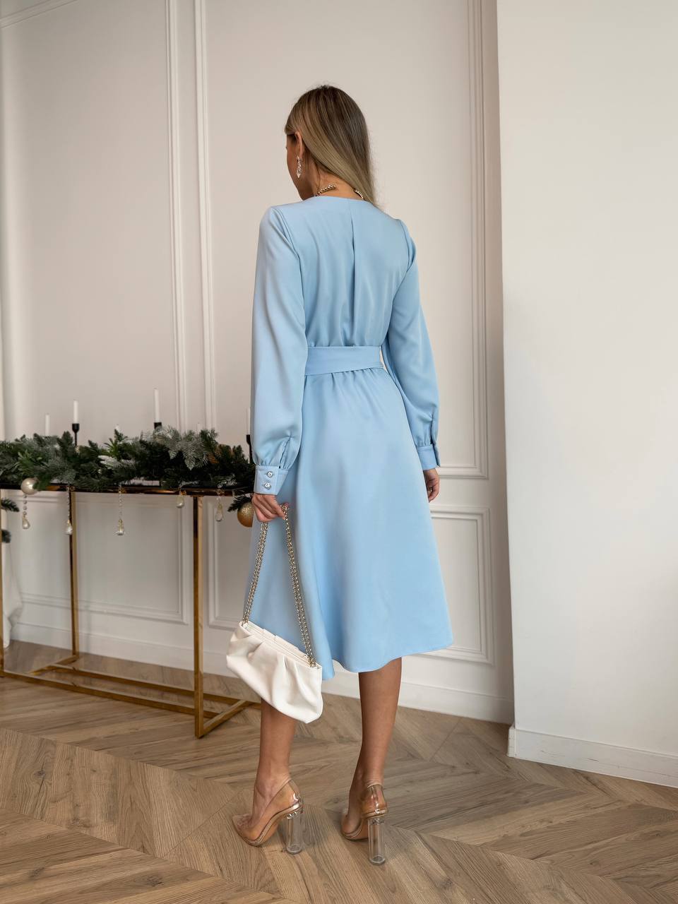 Woman in a light blue dress standing in a room with a decorative table and wall.