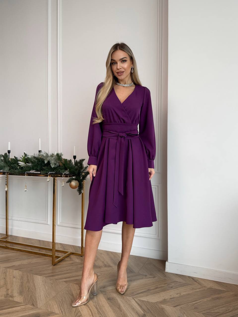 Woman wearing a purple dress standing in a room with a decorative table in the background.