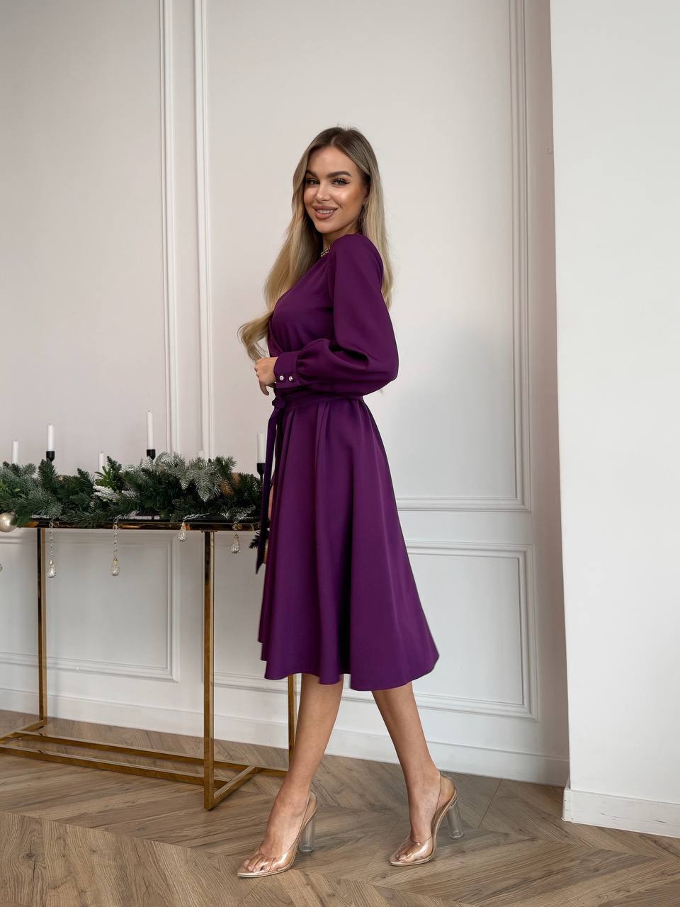Woman in a purple dress standing in a room with a white wall and wooden floor.