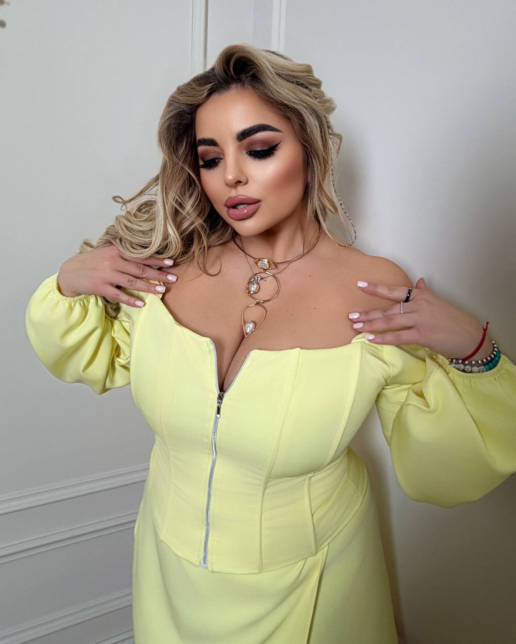Woman wearing a light yellow dress with puffed sleeves against a white wall.