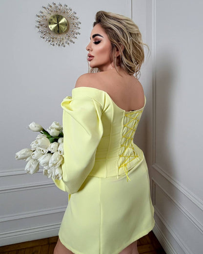 Woman in a yellow off-shoulder dress holding white flowers against a decorative wall.